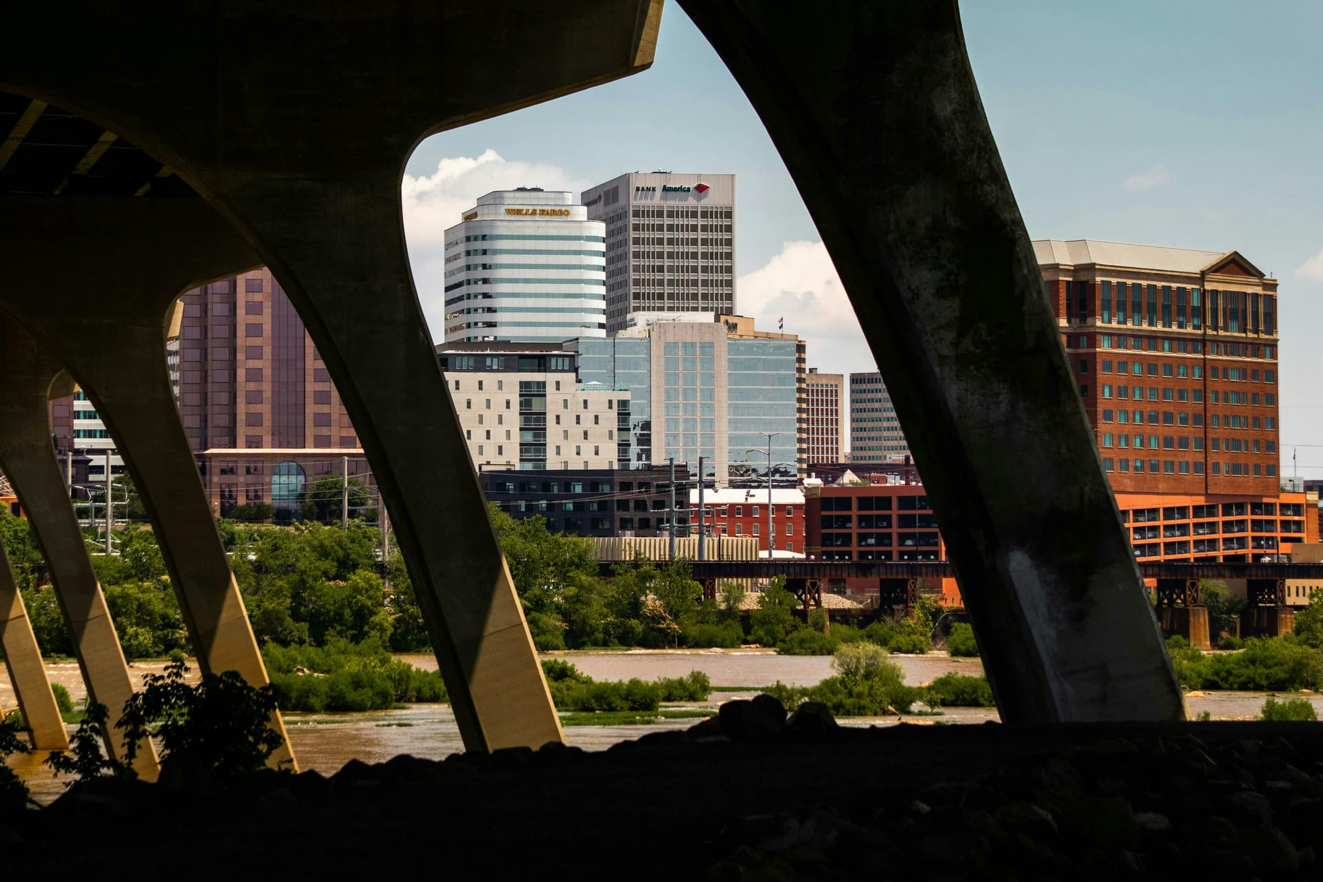 View of a cityscape framed by large concrete arches of a bridge, showcasing a contrast between the modern downtown buildings and the natural riverfront. The buildings include various banks and corporate offices, with a mix of architectural styles visible against a bright blue sky.